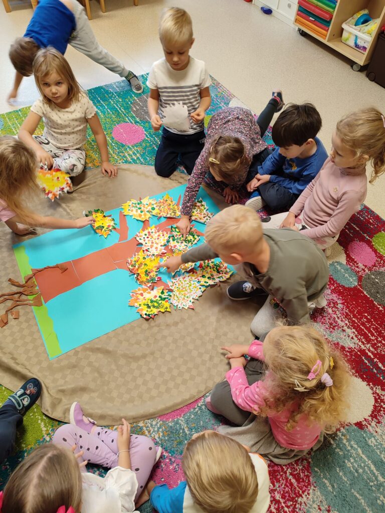 Dzieci przyklejają wykonane liście na sylwetę drzewa.