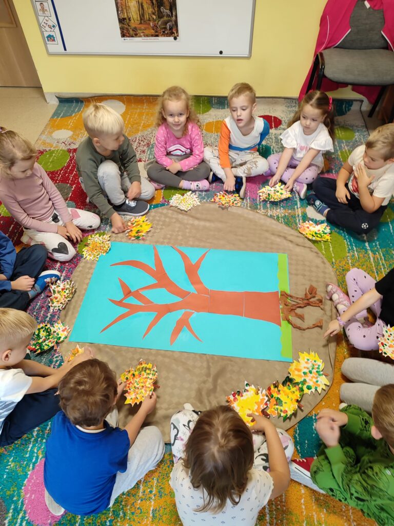 Dzieci przyklejają wykonane liście na sylwetę drzewa.