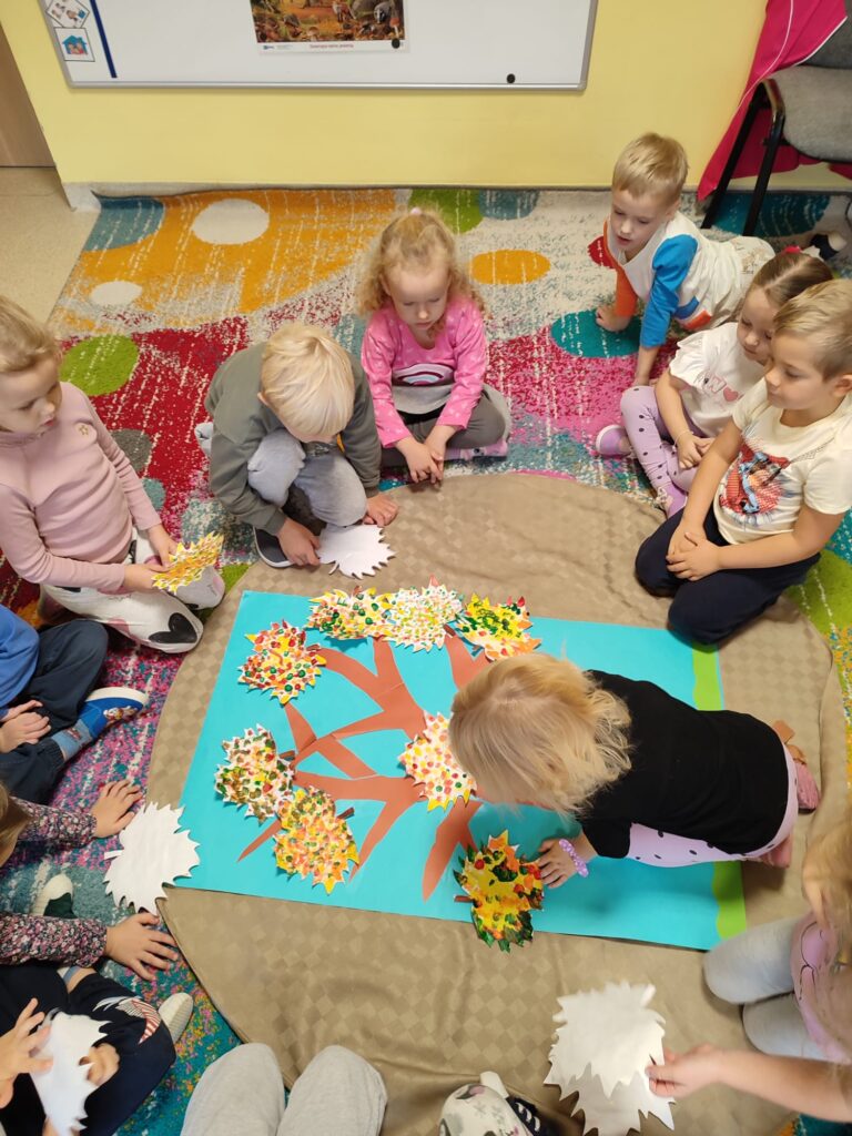 Dzieci przyklejają wykonane liście na sylwetę drzewa.