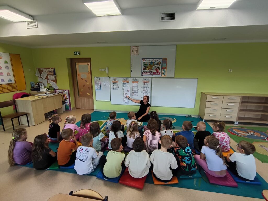 Przedszkolaki siedzą na dywanie słuchają nauczyciela.
